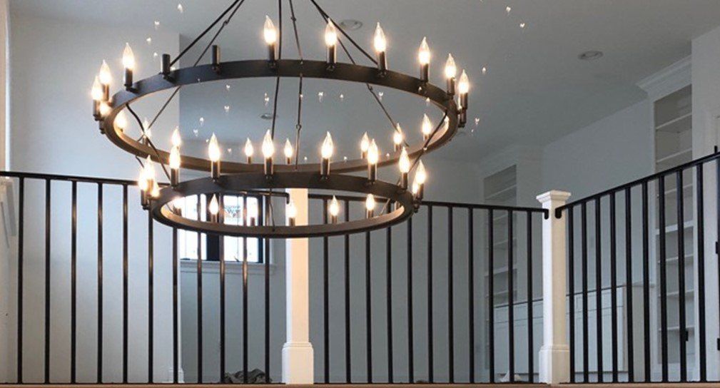 Two-tiered black metal chandelier hangs in a room with white walls, black railing, and a light-colored floor.