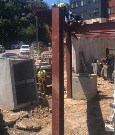 Construction workers on a site welding steel beams. Concrete structures and parked vehicles visible.