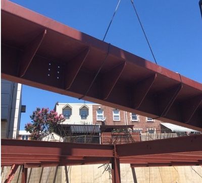 A red steel beam being lifted by a crane at a construction site with buildings in the background.