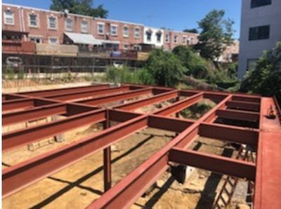 Red steel beams form the foundation of a building on a dirt lot. Row houses are in the background.