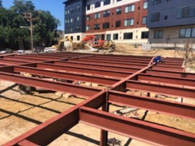 Steel beams in a grid, likely the foundation of a building under construction, in front of a multi-story building.