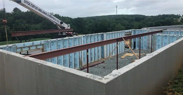 Construction site with crane lifting steel beams over a concrete and blue foam block foundation.
