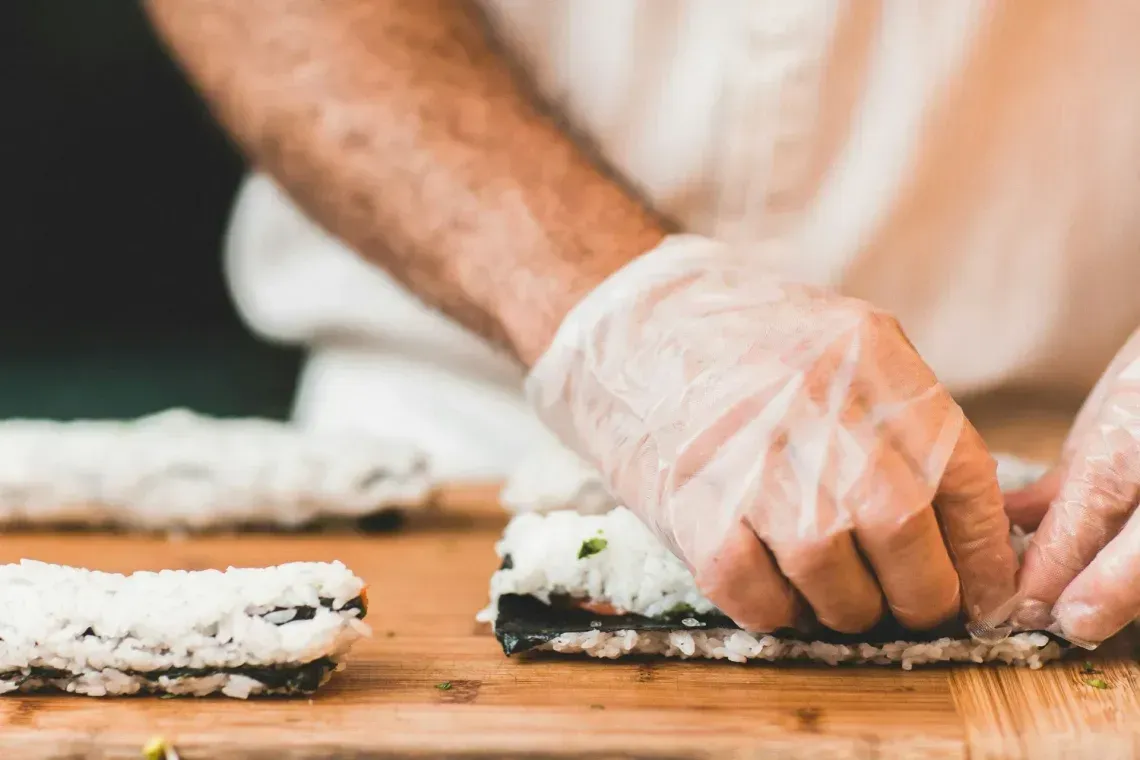 Gloved hands shaping sushi rolls on a wooden cutting board