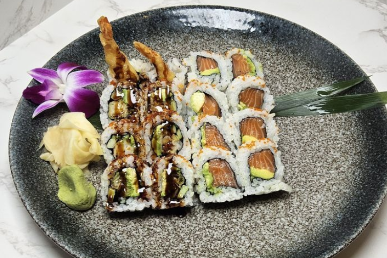 Sushi rolls on a black plate, topped with sauce, garnished with wasabi, ginger, and a purple orchid.