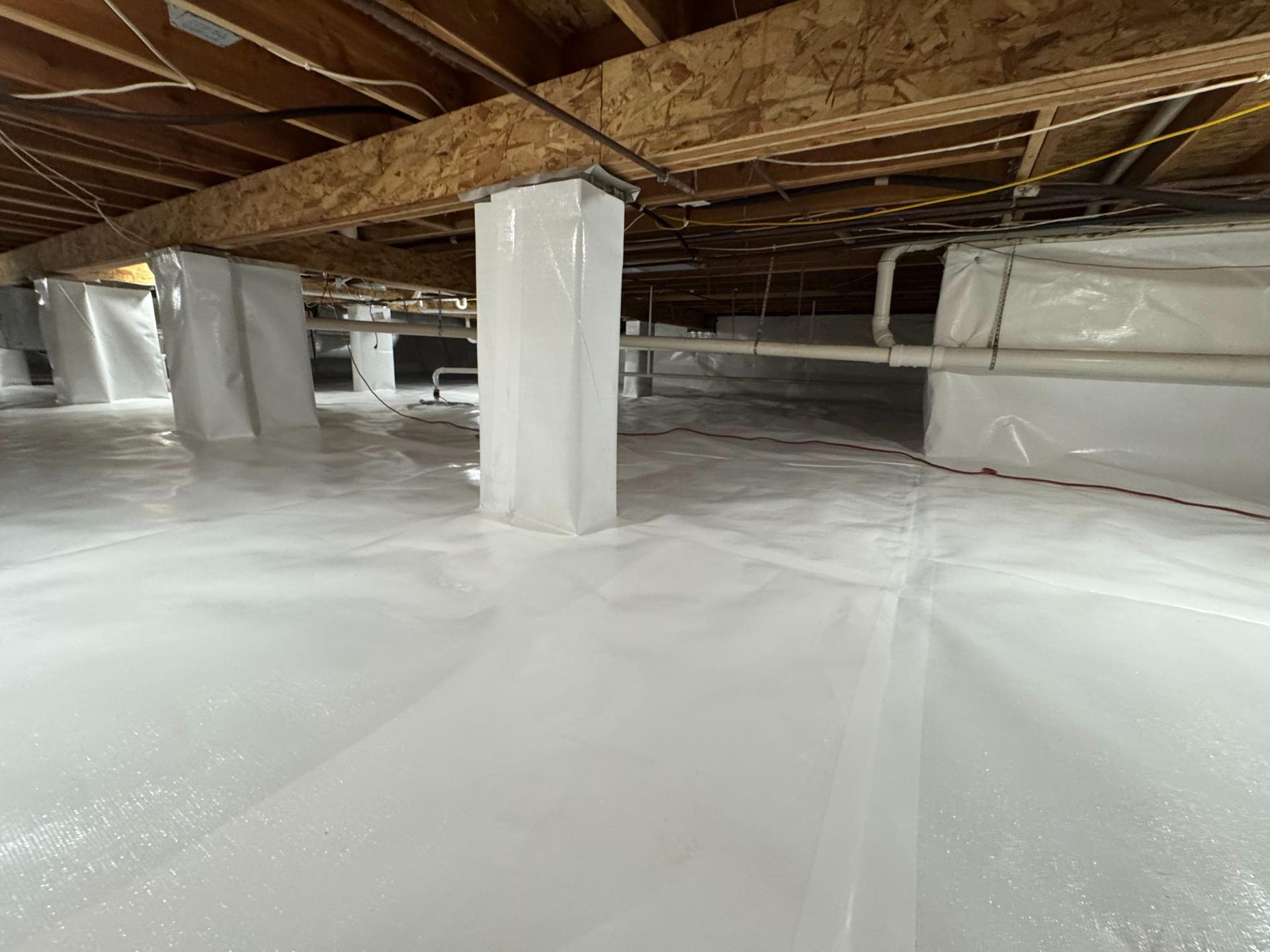 A crawl space with white plastic vapor barrier covering the ground and wrapping around the support pillars and walls.