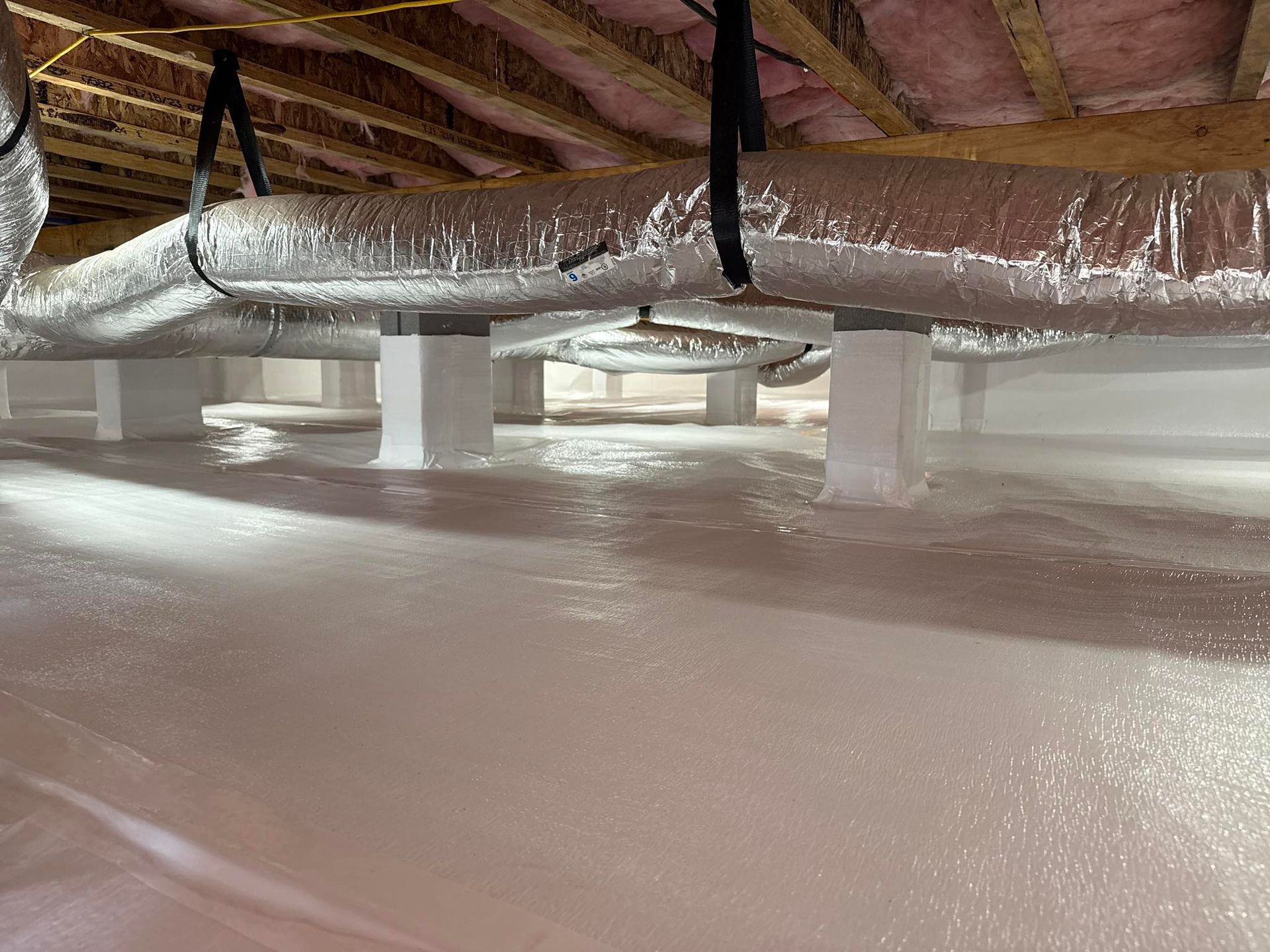 Crawl space with insulated HVAC ducts and support pillars resting on a white vapor barrier covering the ground.