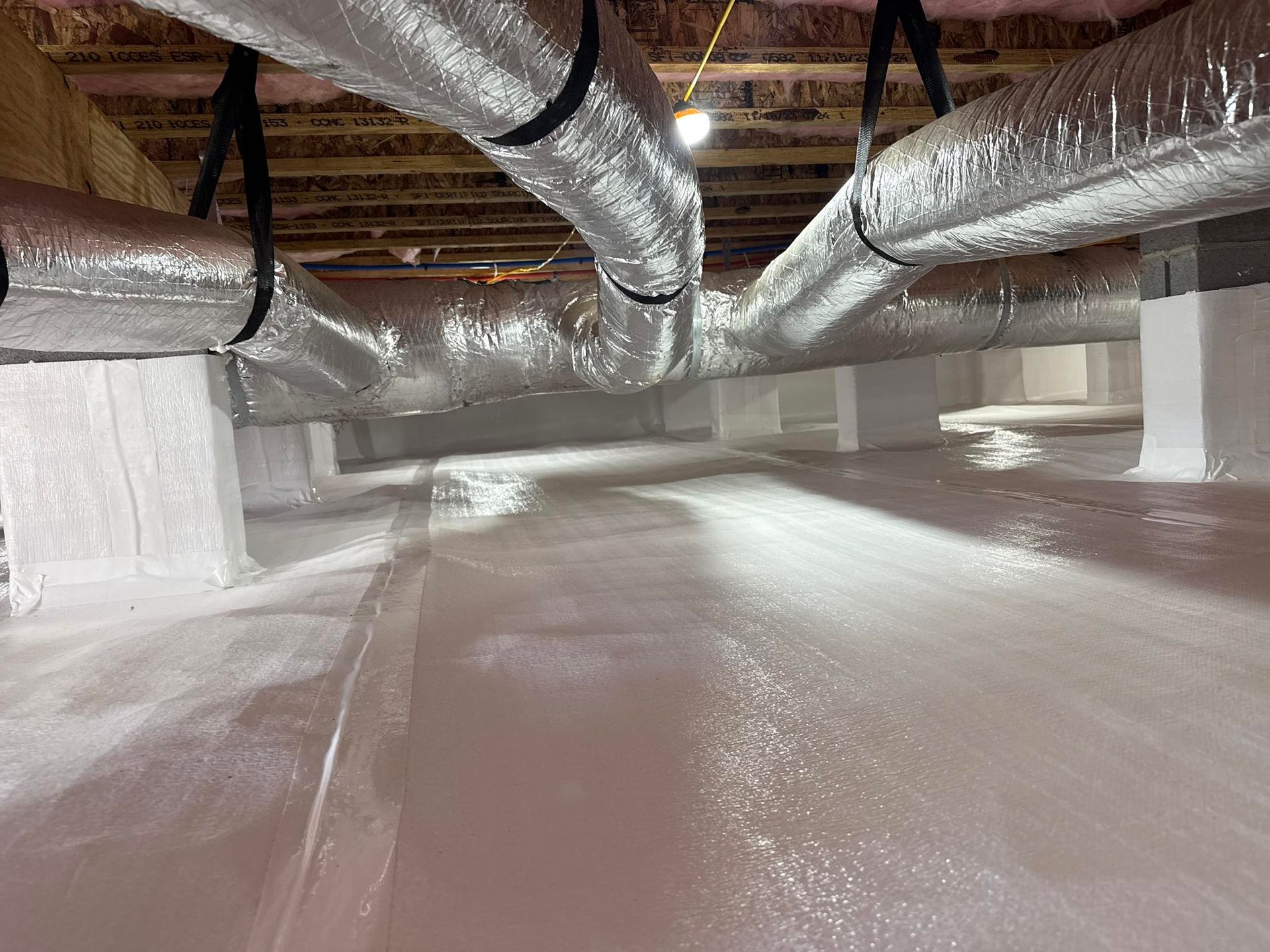 A crawl space with silver insulated ductwork suspended from wooden ceiling joists above a bright white vapor barrier.