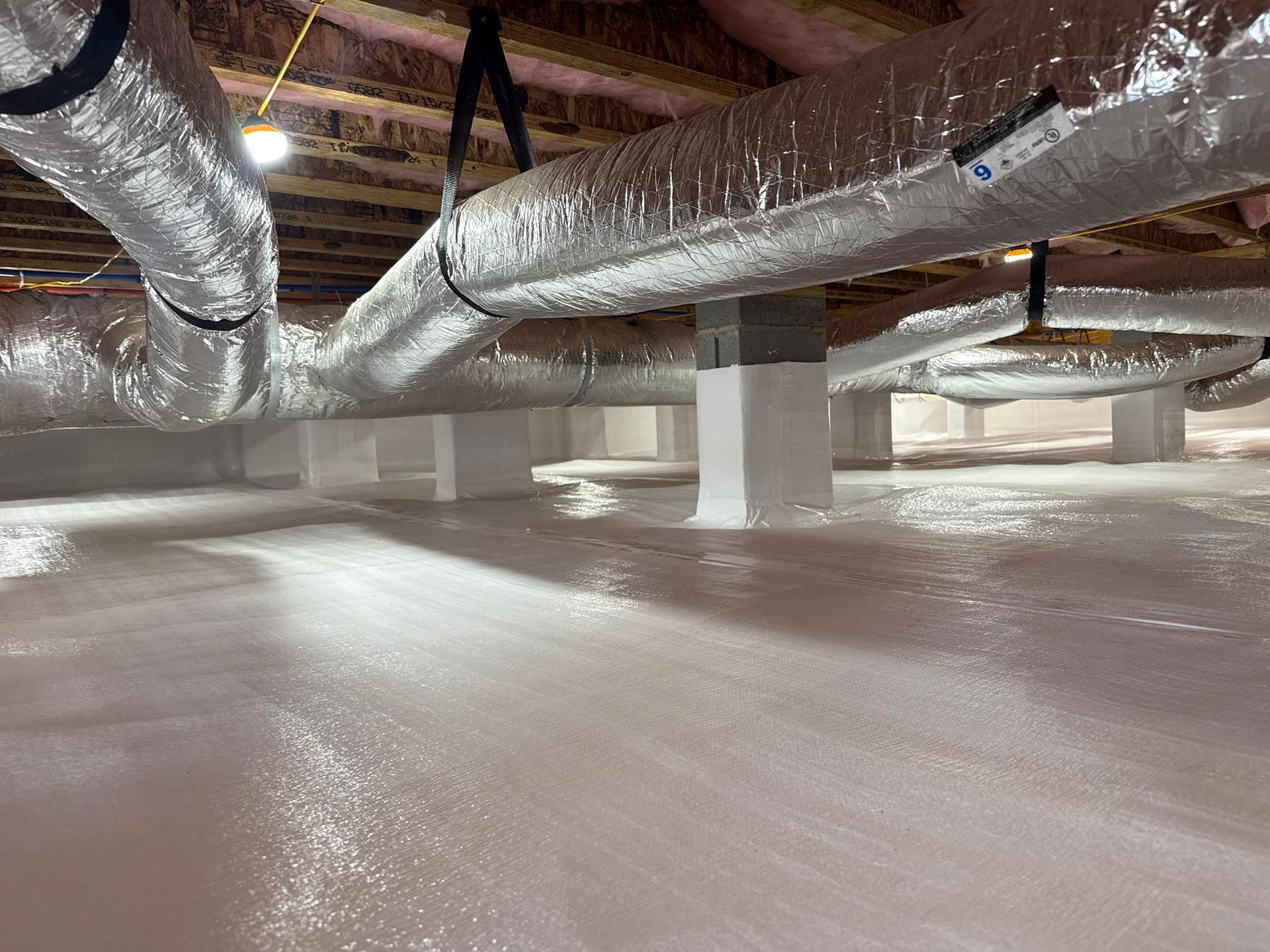 A clean crawl space with a vapor barrier floor, support columns, and insulated silver HVAC ducts suspended overhead.