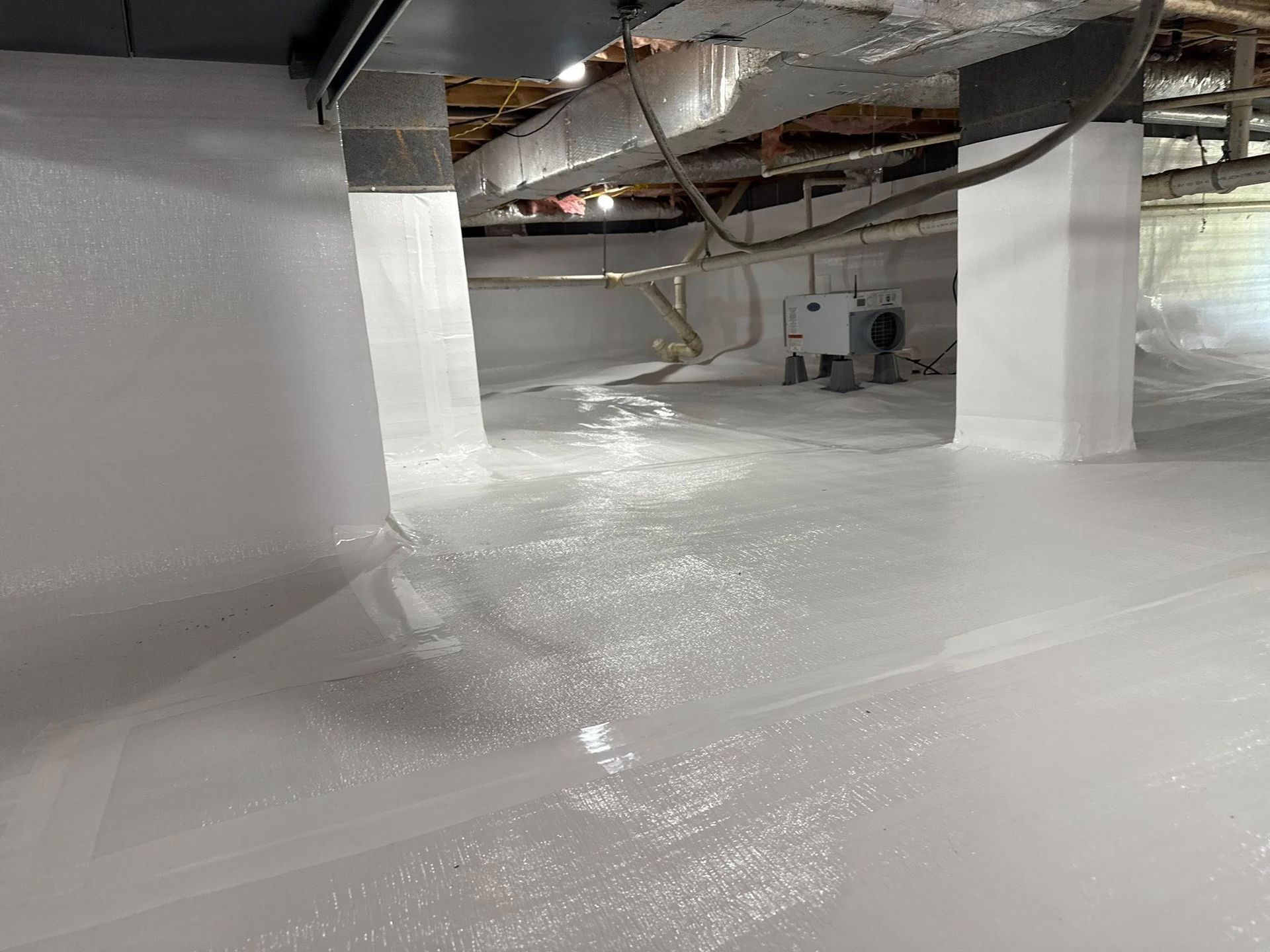 A crawl space encapsulated with white vapor barrier plastic on the floor and walls, with a dehumidifier in the background.