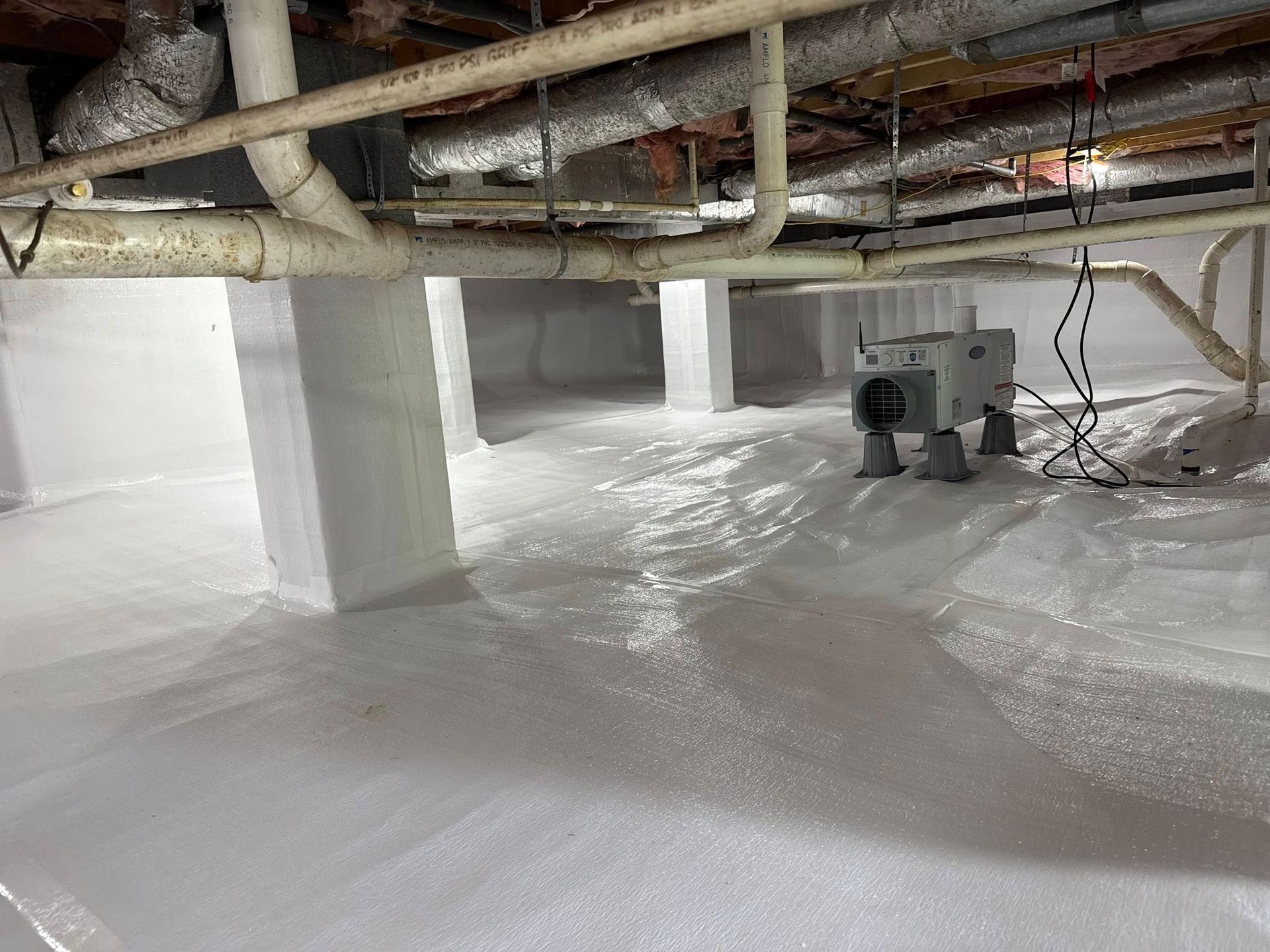 A crawl space encapsulated with white vapor barrier, featuring support columns, exposed ceiling pipes, and a dehumidifier.