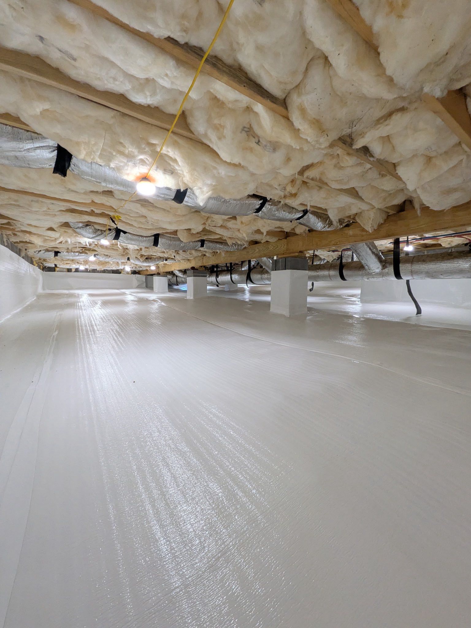 A crawl space with a white vapor barrier on the ground and fiberglass insulation tucked between the wooden floor joists.