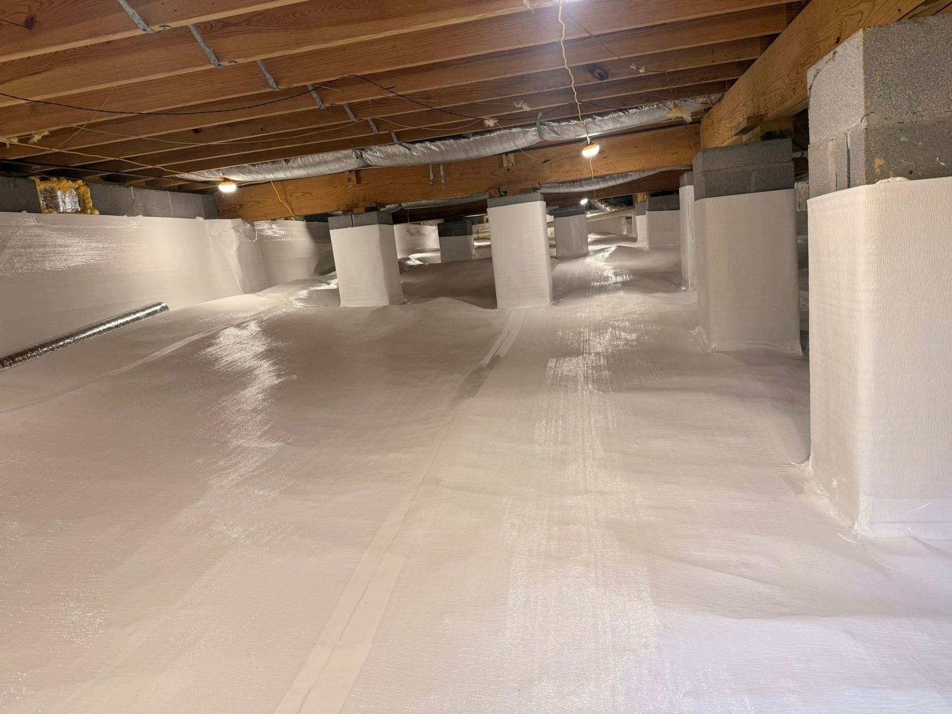 A crawl space with white moisture barrier lining the floor and wrapping around support pillars under wooden floor joists.