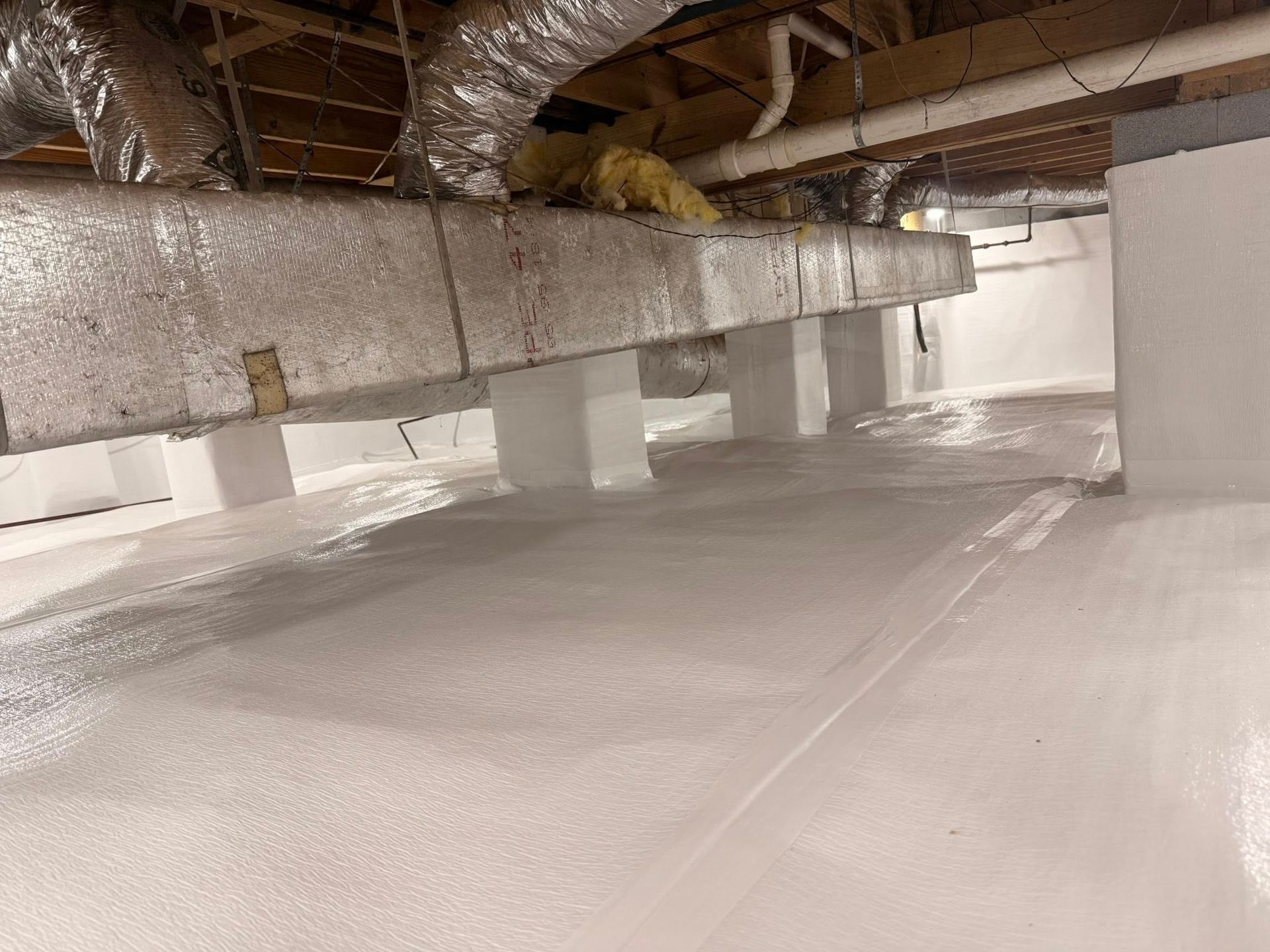 A crawl space with white encapsulation on the walls and floor, featuring support piers and silver ductwork above.