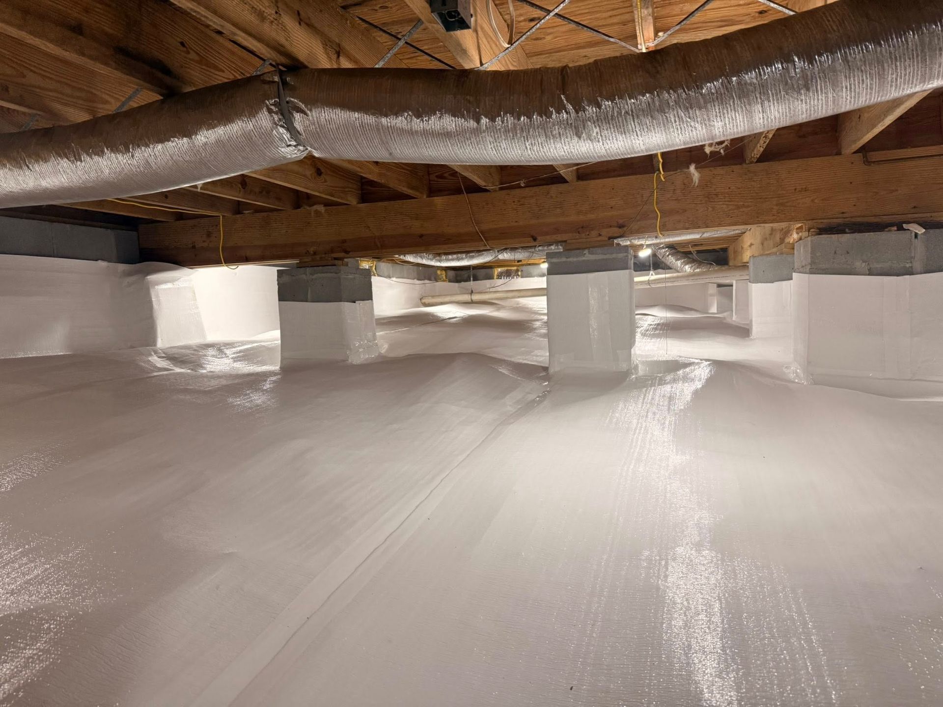A crawl space with white moisture barrier lining the ground and pillars, and wooden joists and ductwork on the ceiling.