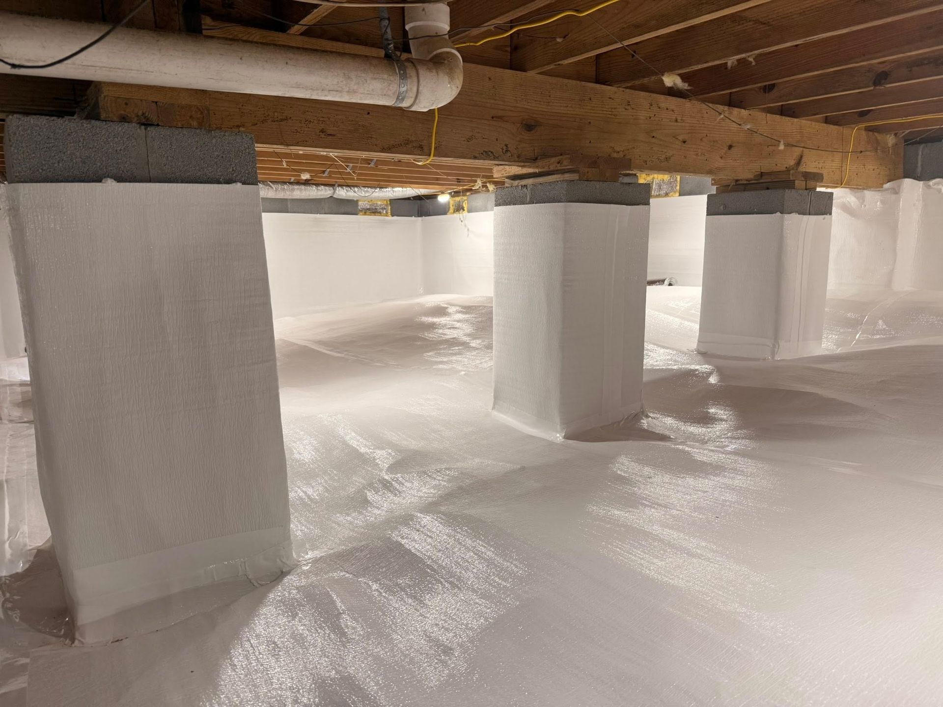 A crawl space with concrete support pillars wrapped in white vapor barrier material and a white ground liner.