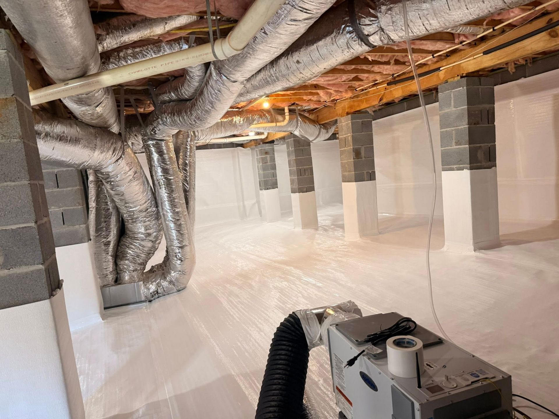 A crawlspace with insulated silver ductwork, white vapor barrier flooring, and a dehumidifier in the foreground.