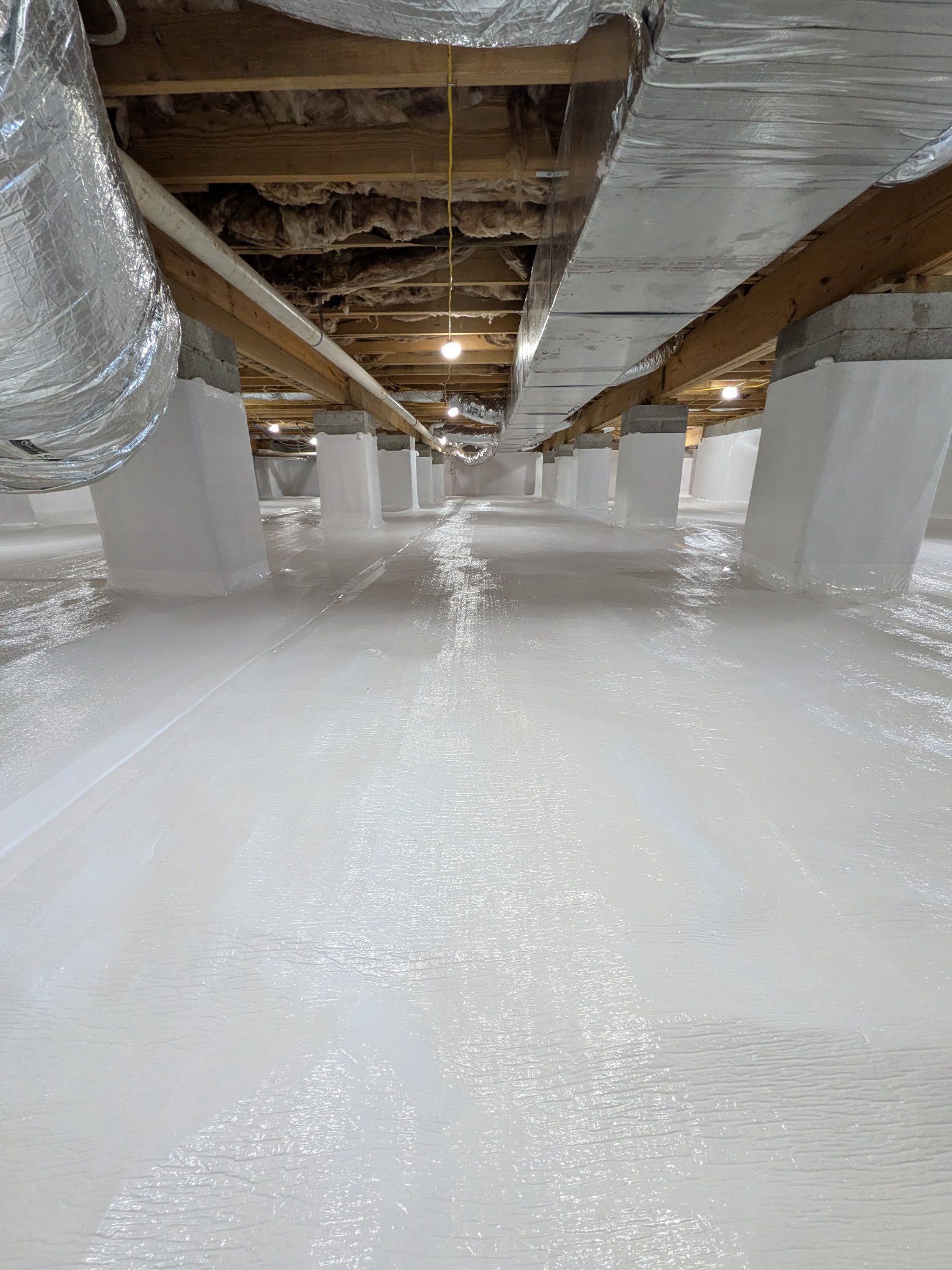 Crawl space featuring white vapor barrier on the floor and piers, with insulated ductwork attached to wooden floor joists.