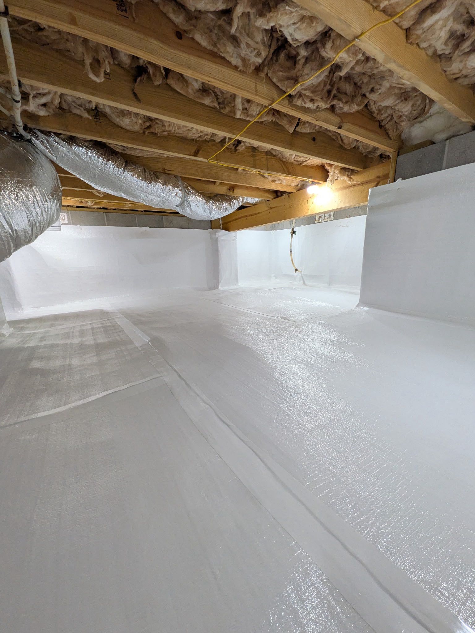 A crawl space with white vapor barrier flooring and walls, and fiberglass insulation between the wooden ceiling joists.