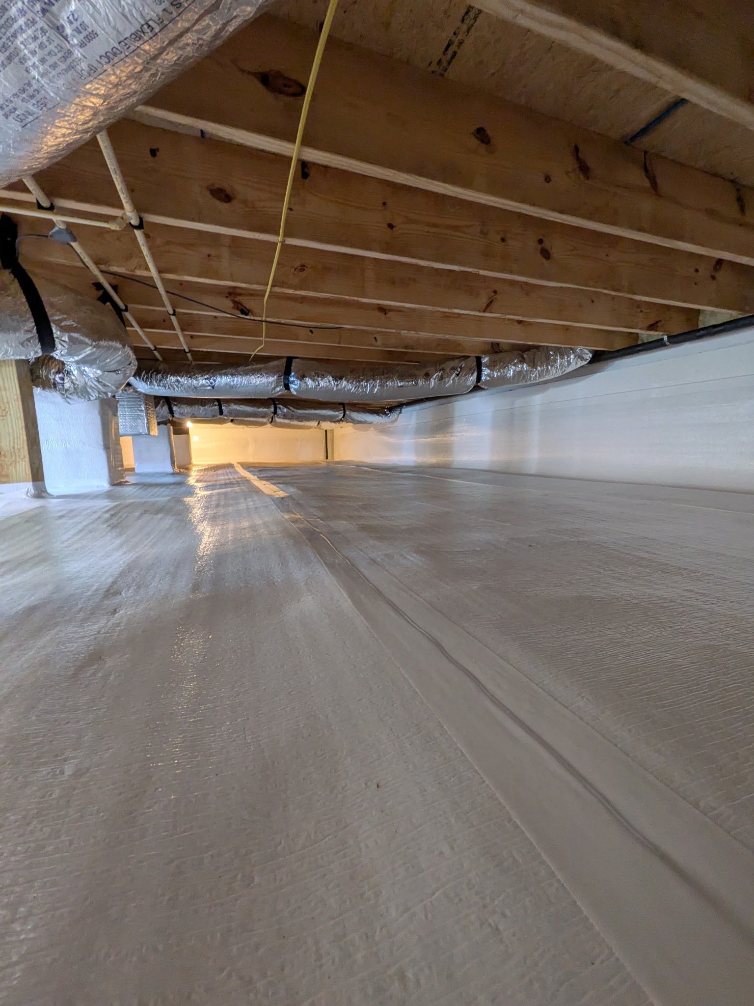 A low-angle view of a crawl space with exposed wooden floor joists, reflective insulation, and a clean, sealed vapor barrier.
