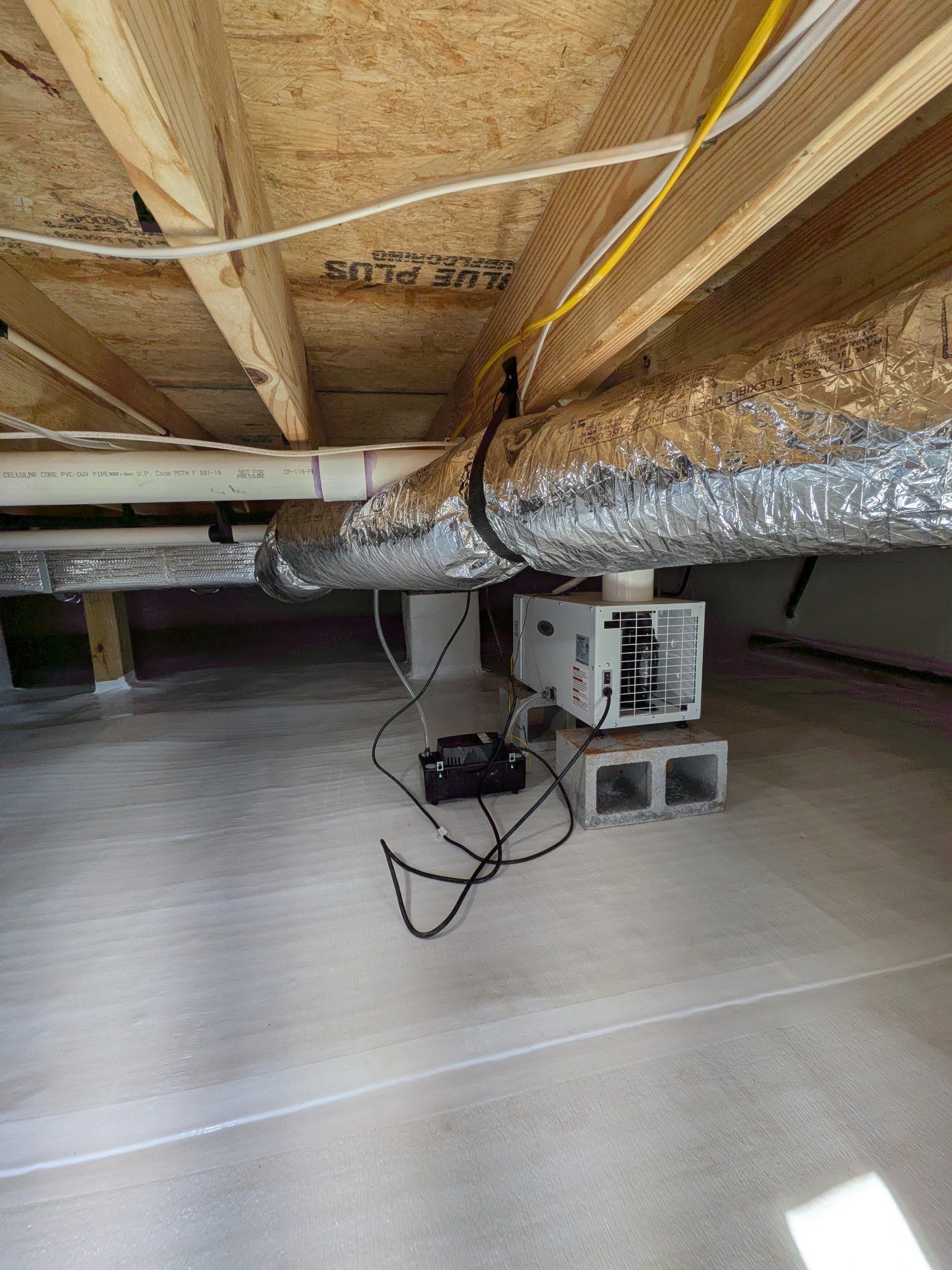 A white crawlspace dehumidifier sits on concrete blocks in a clean, encapsulated crawlspace with exposed joists above.