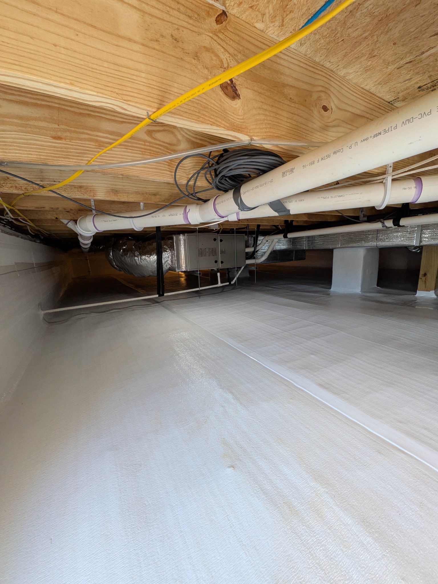 A crawl space with a clean white vapor barrier, exposed wooden floor joists, white PVC pipes, and a metal HVAC duct.