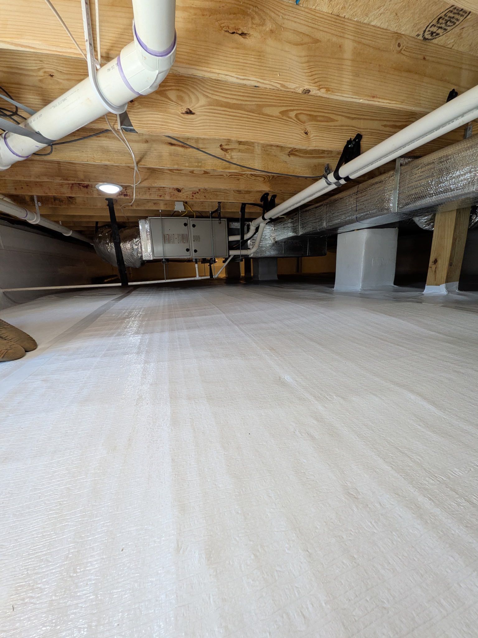 A view of a crawl space with a vapor barrier on the ground, overhead floor joists, white PVC plumbing, and HVAC ducts.