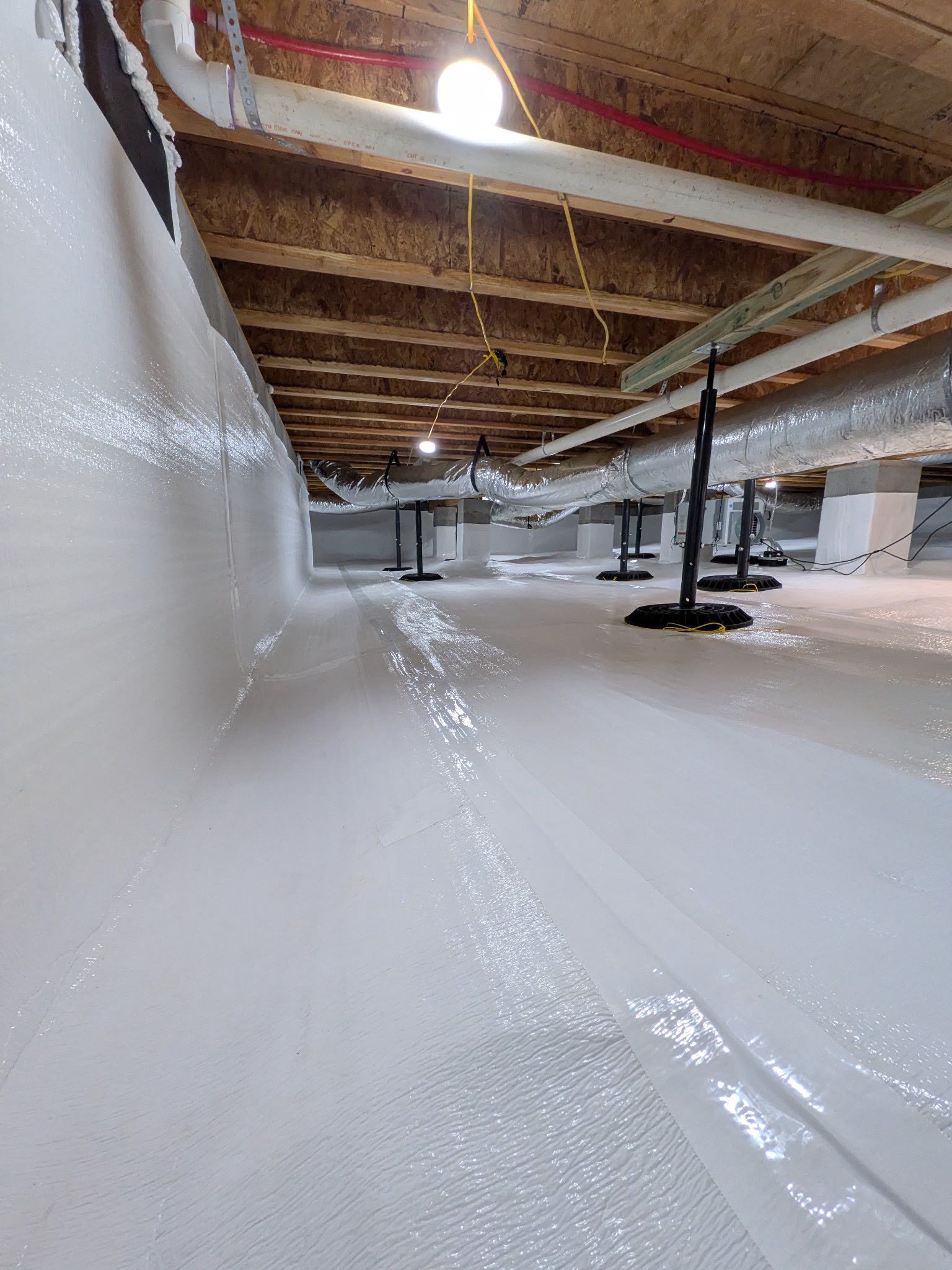 A crawl space with white moisture-barrier lining on the walls and floor, wooden joists above, and black support pillars.