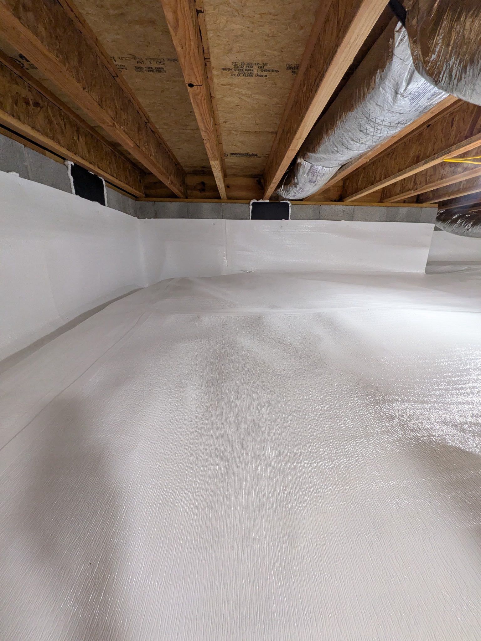 A white vapor barrier installed in a crawl space, showing sealed foundation walls and exposed wooden floor joists above.