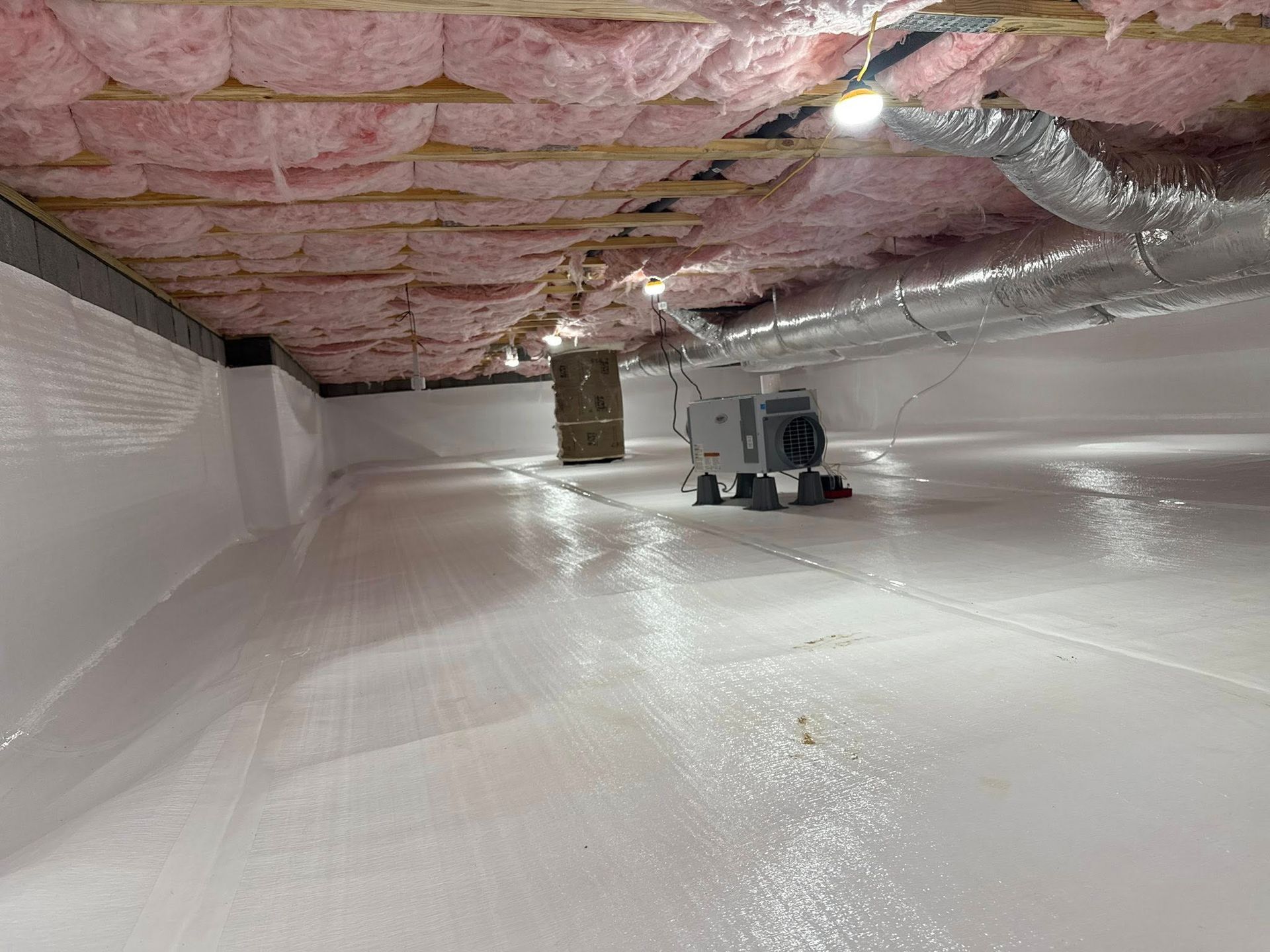 A crawlspace with a white moisture barrier on the floor and walls, pink fiberglass insulation, and a dehumidifier.
