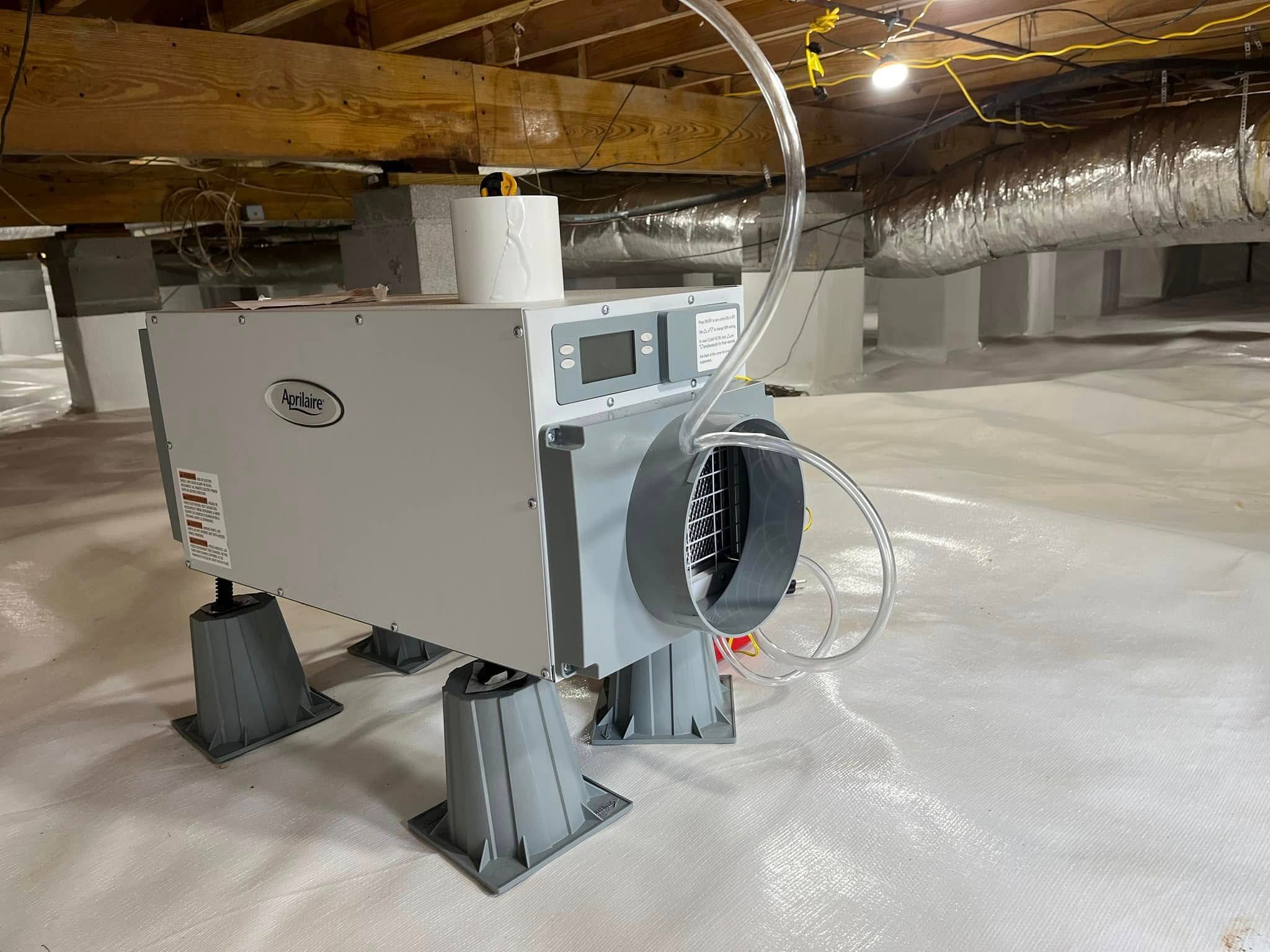 A crawl space dehumidifier stands on plastic risers atop a vapor barrier in a dimly lit, unfinished crawl space.