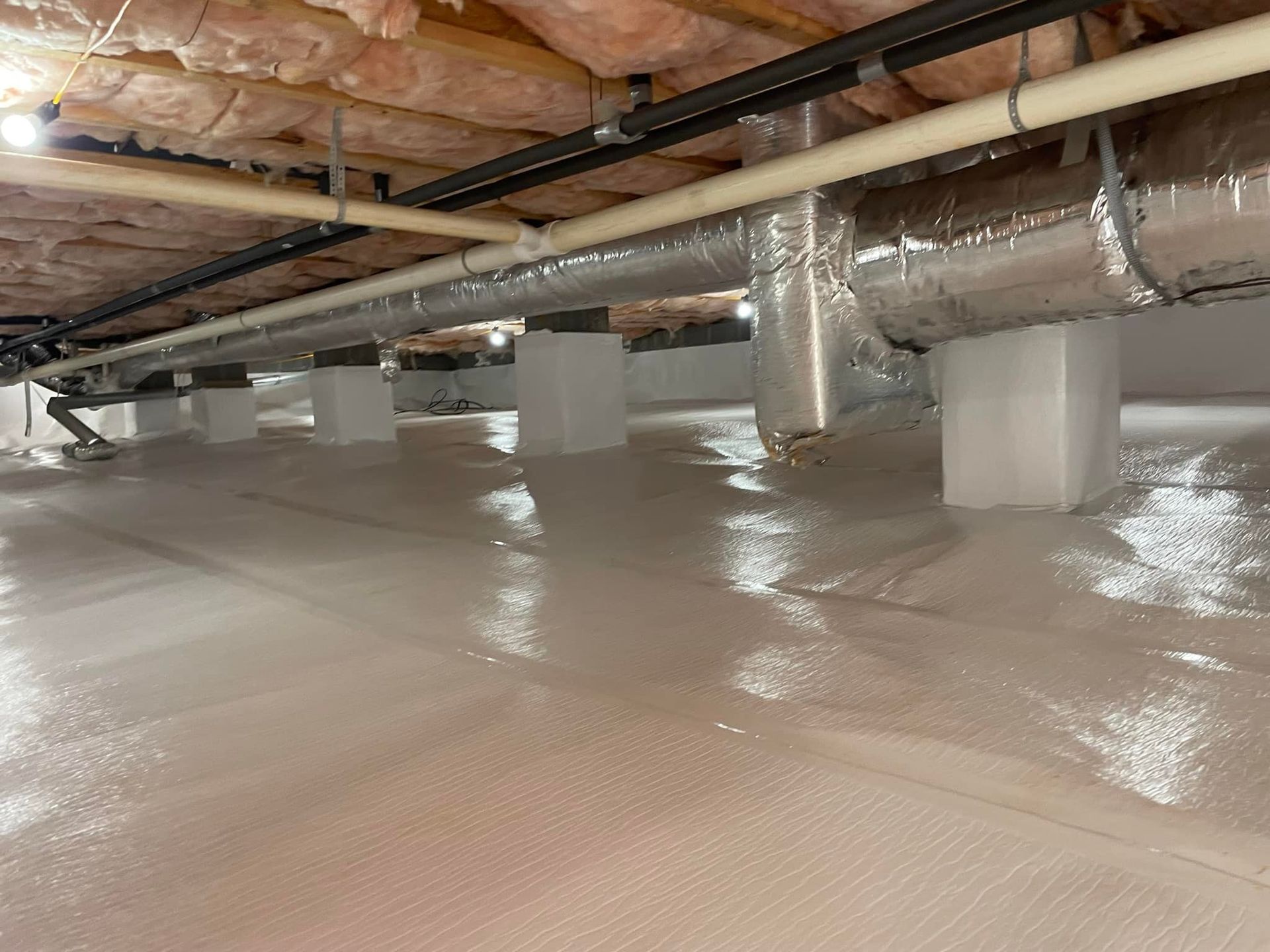 A clean, well-lit crawl space with a white vapor barrier on the ground, concrete support pillars, and overhead ductwork.