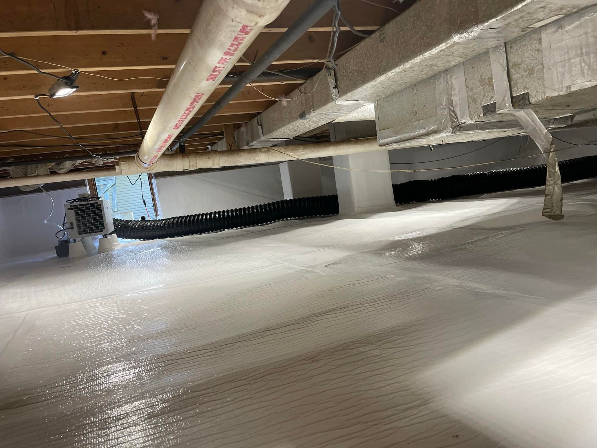 A clean crawl space with a bright white vapor barrier covering the floor and a dehumidifier in the background.