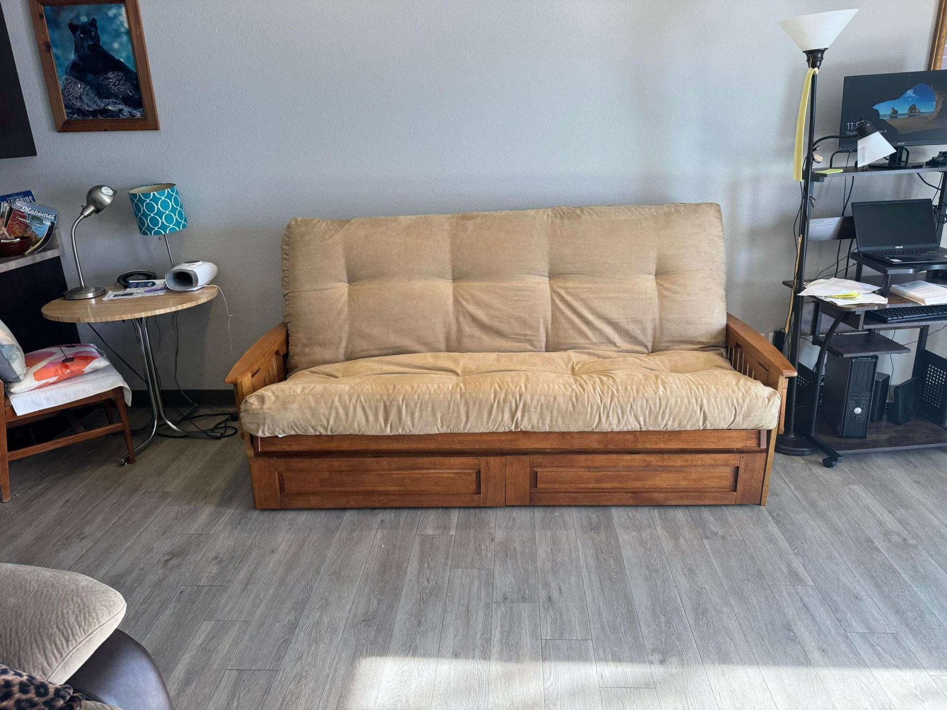 A tan futon with a wooden frame sits centered in a room with light flooring, a side table, and a desk nearby.