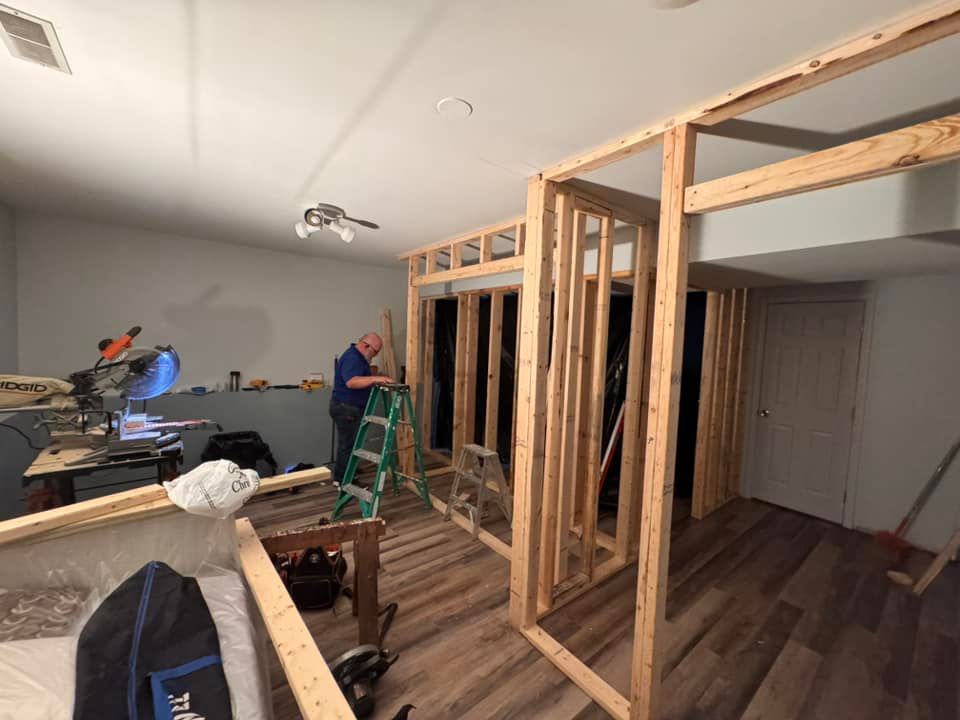 A person stands on a ladder working on a wood-framed wall in a room with light grey walls and wood-patterned flooring.