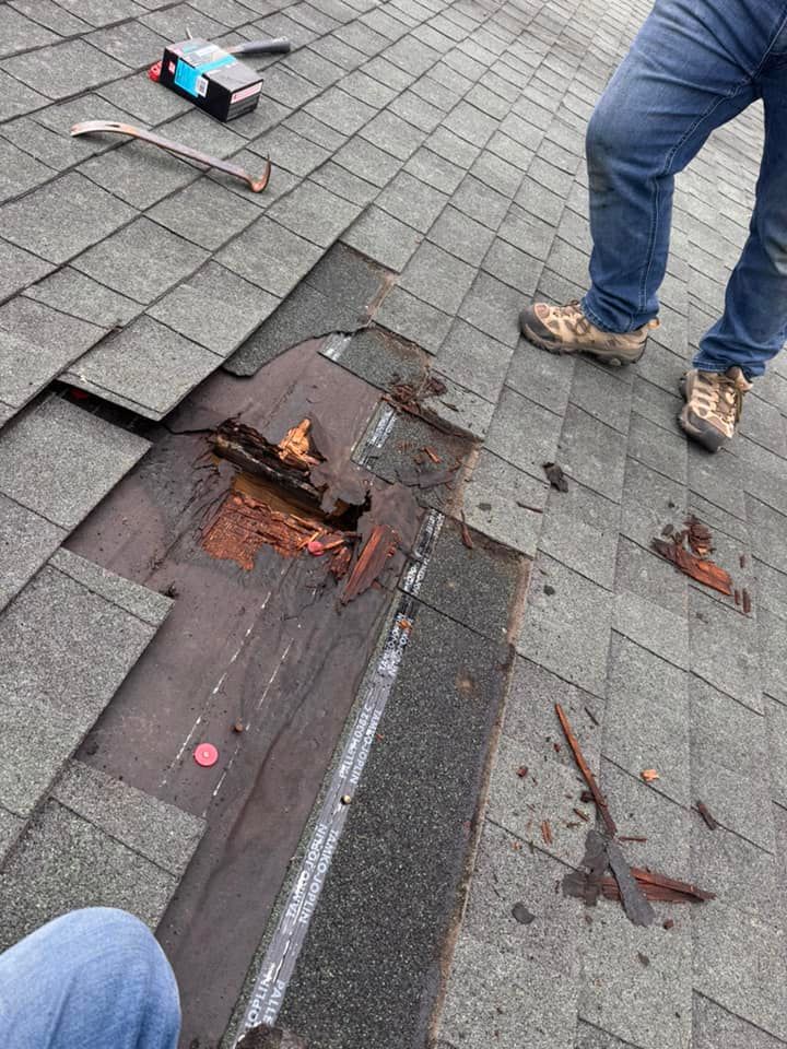 A person stands on a roof next to a hole revealing rotted wooden decking and roofing felt, with tools nearby.