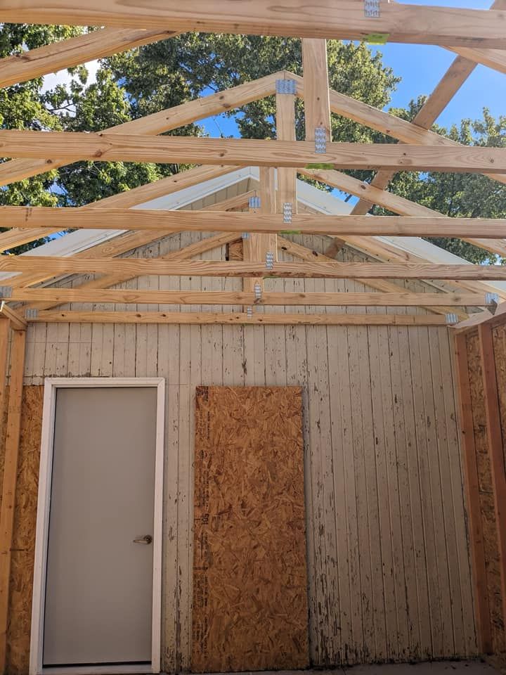 A new wooden roof truss structure under construction above a partially boarded wall with a single white door.