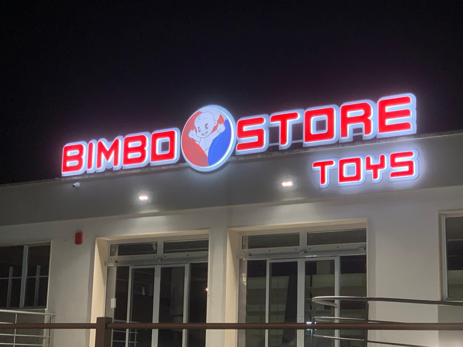 "Insegna di Bimbo Store Toys, lettere rosse, logo a fumetti, sopra un negozio con porte scorrevoli in vetro, impostazione notturna."