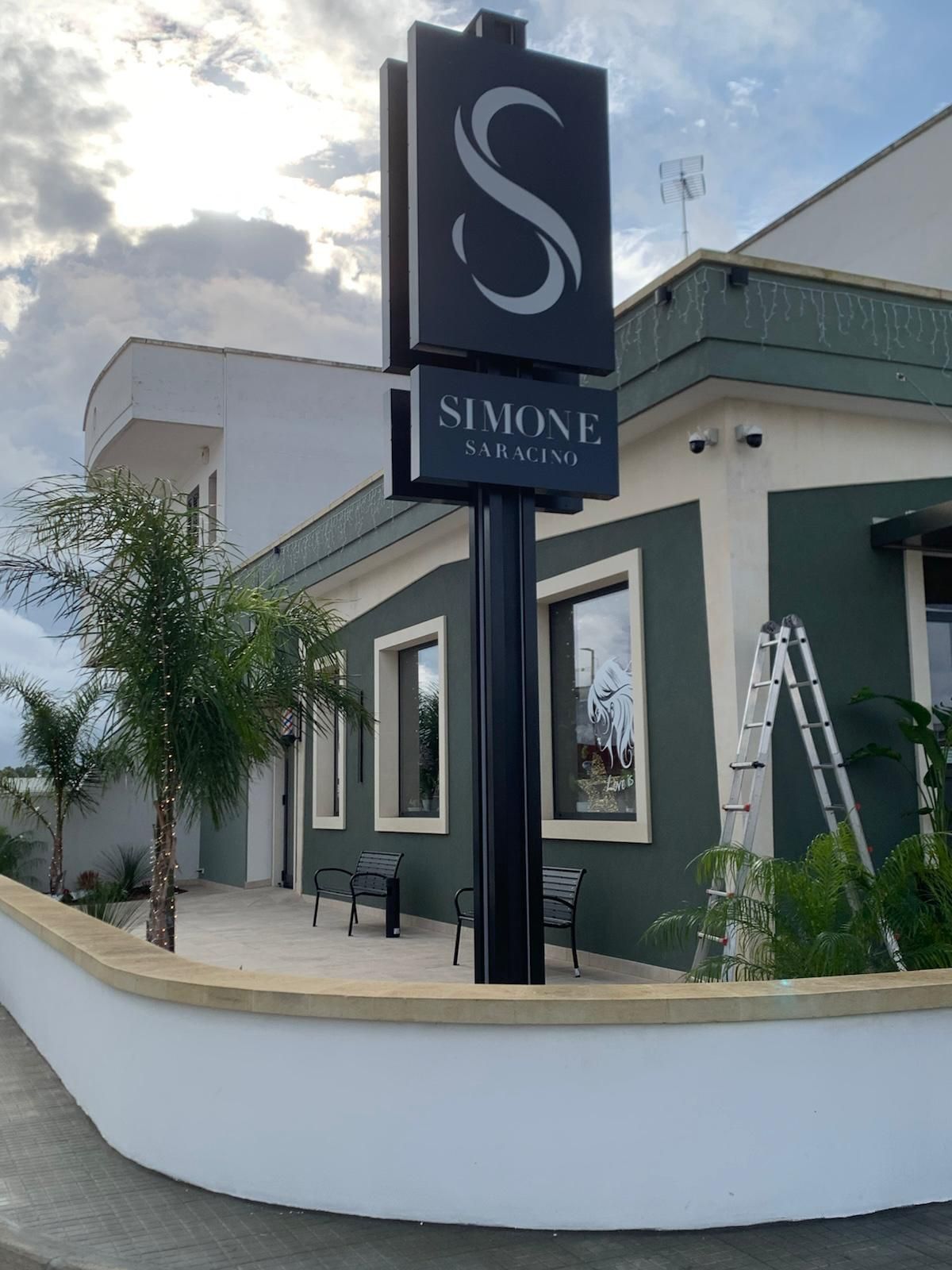 Cartello per "Simone Swimming" con grande logo "S" nero, montato su un palo nero all'esterno di un edificio con facciata verde e bianca.