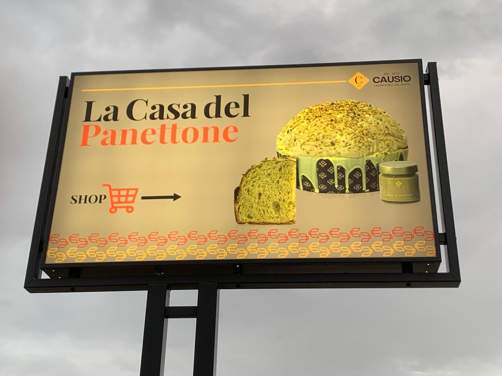 Cartellone pubblicitario per "La Casa del Panettone" con torta e barattolo di panettone, icona del negozio, design arancione e giallo.
