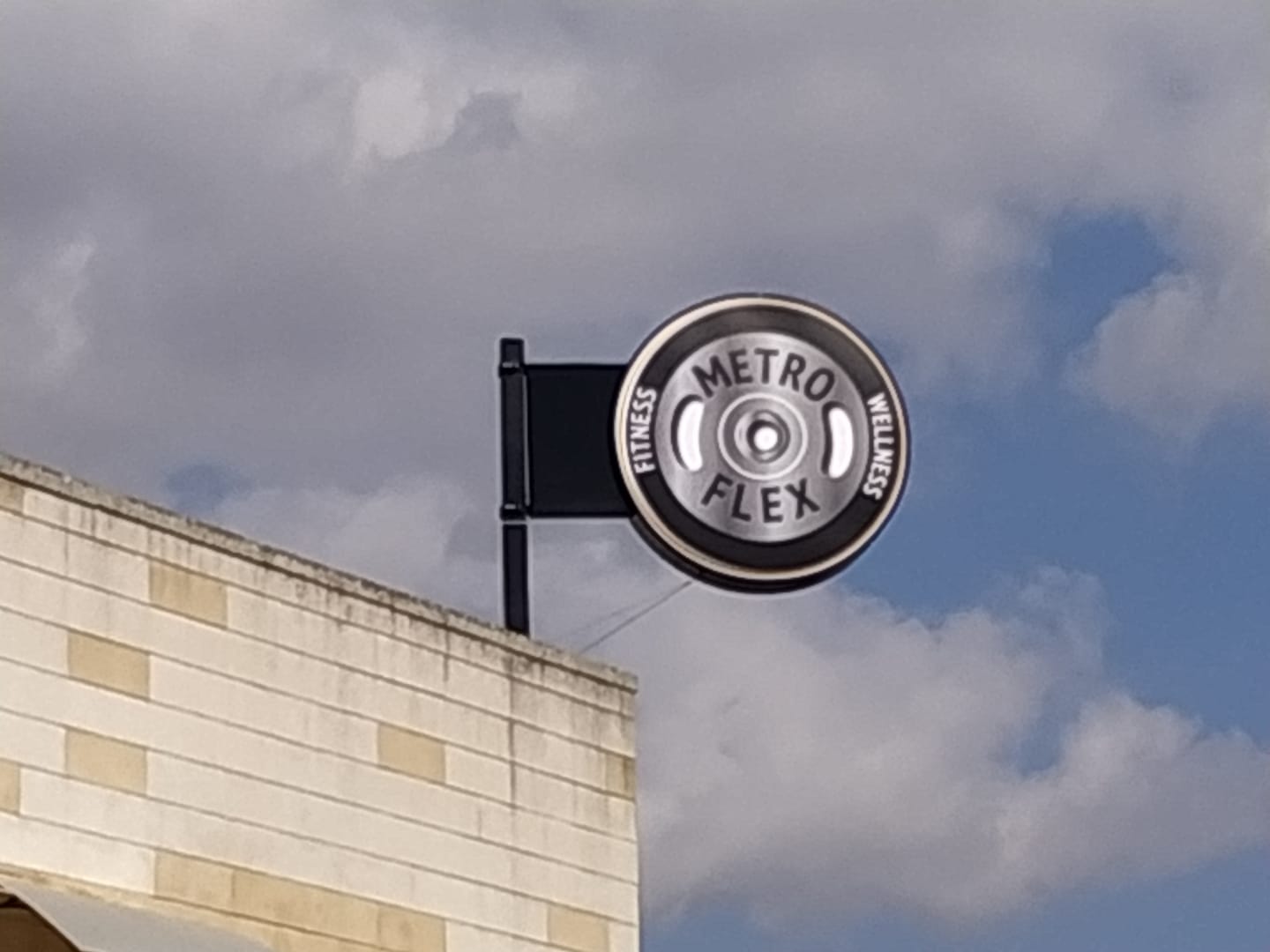 Cartello per il centro fitness Metro Flex, un disegno circolare che ricorda un peso, attaccato a un edificio sullo sfondo di un cielo nuvoloso.