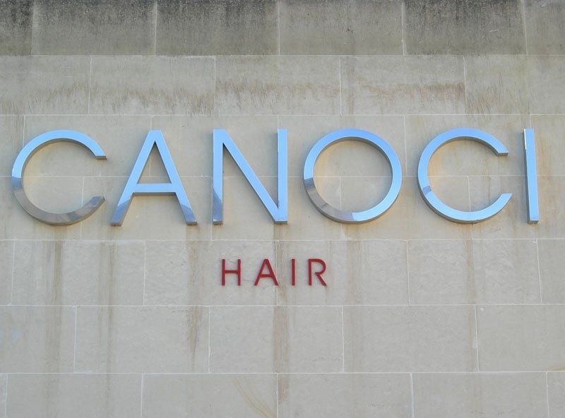 "CANOCI" in lettere argentate sopra "HAIR" in rosso, su una parete beige chiaro.