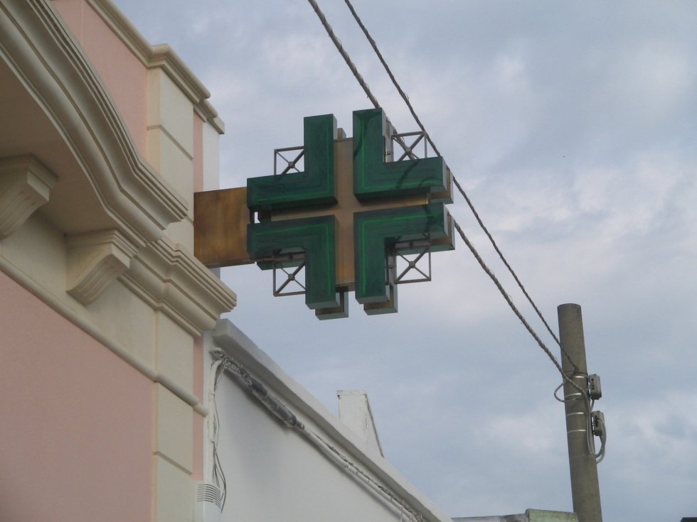 Cartello di farmacia con croce verde attaccato a un edificio, con linee elettriche sullo sfondo.
