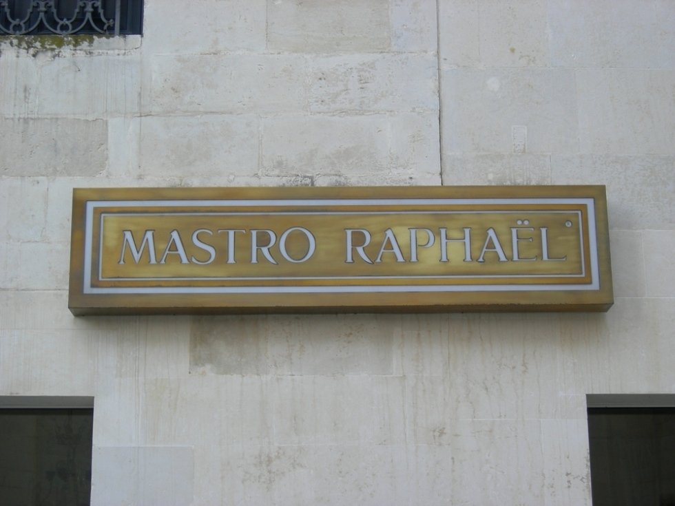 Segnaletica: "MASTRO RAPHAEL" in lettere argentate su una targa con cornice dorata montata su un muro di pietra color crema.