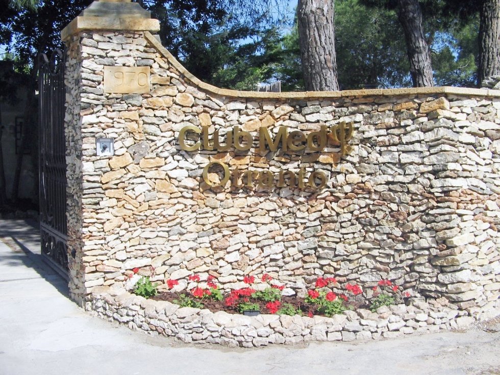 Ingresso con muro in pietra con insegna "Club Mediteranee" e fiori rossi.