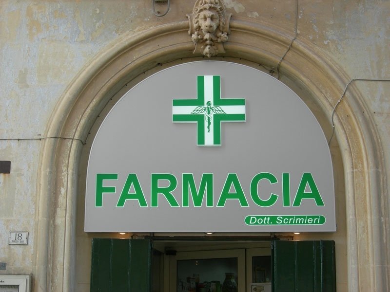Vetrina della farmacia con il simbolo della croce verde e l'insegna "FARMACIA" sopra l'ingresso.