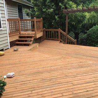 Wooden backyard deck with stairs, railing, and pergola beside a house and greenery.