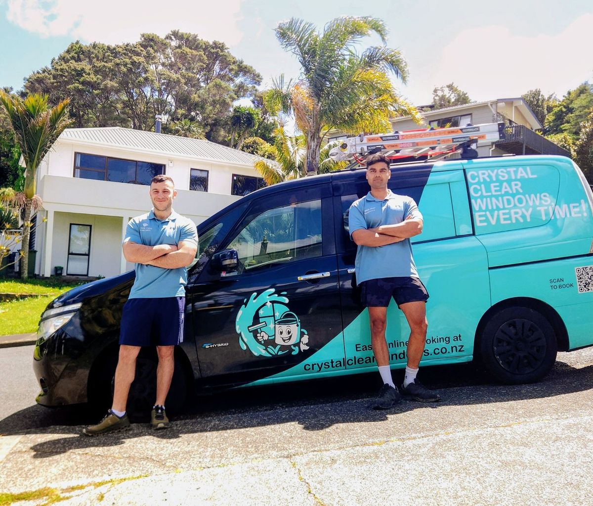 Two men in matching shirts stand by a cleaning van, holding equipment. Black van with teal logo in residential setting.