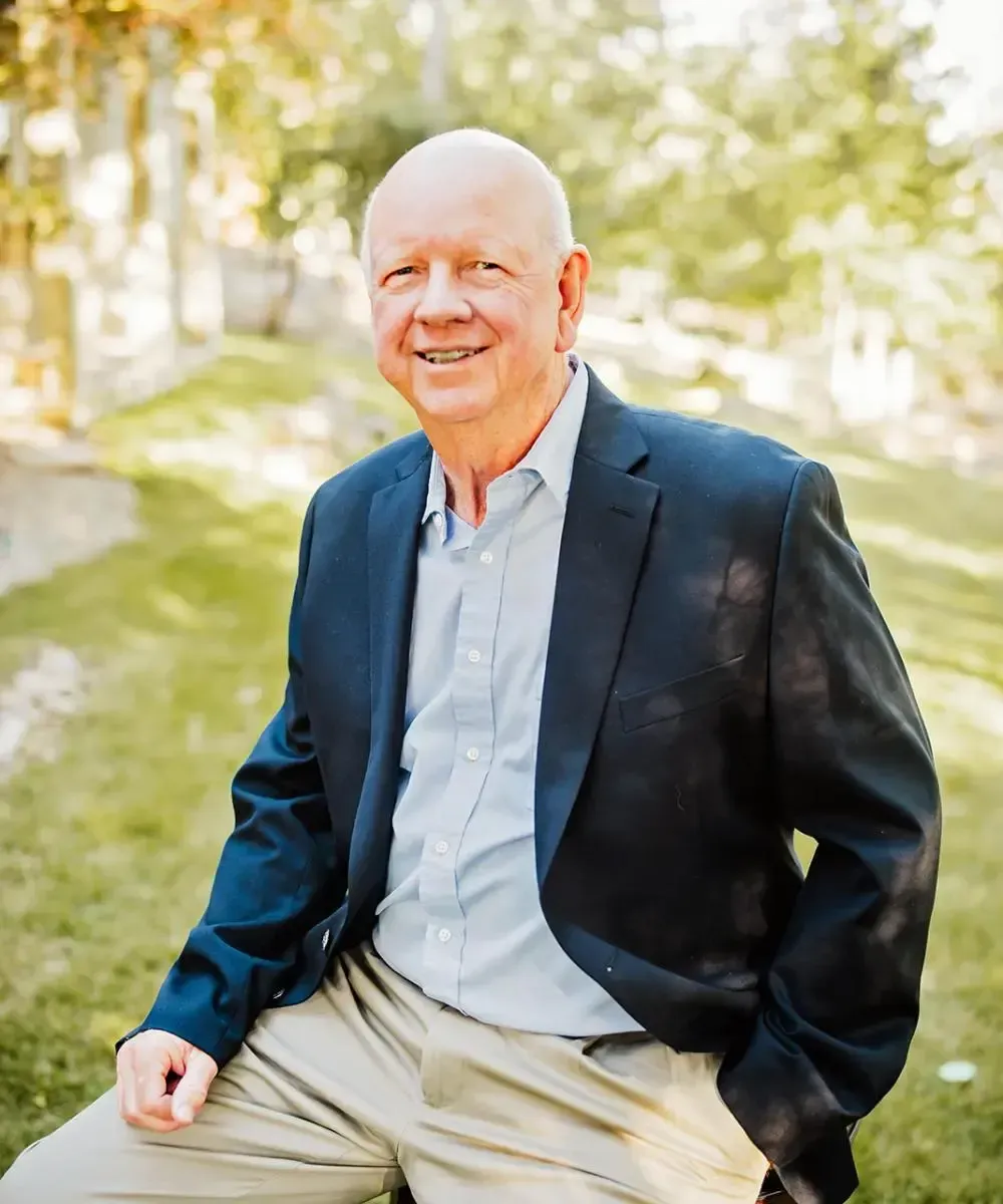 A man in a suit is sitting in the grass with his hands in his pockets.