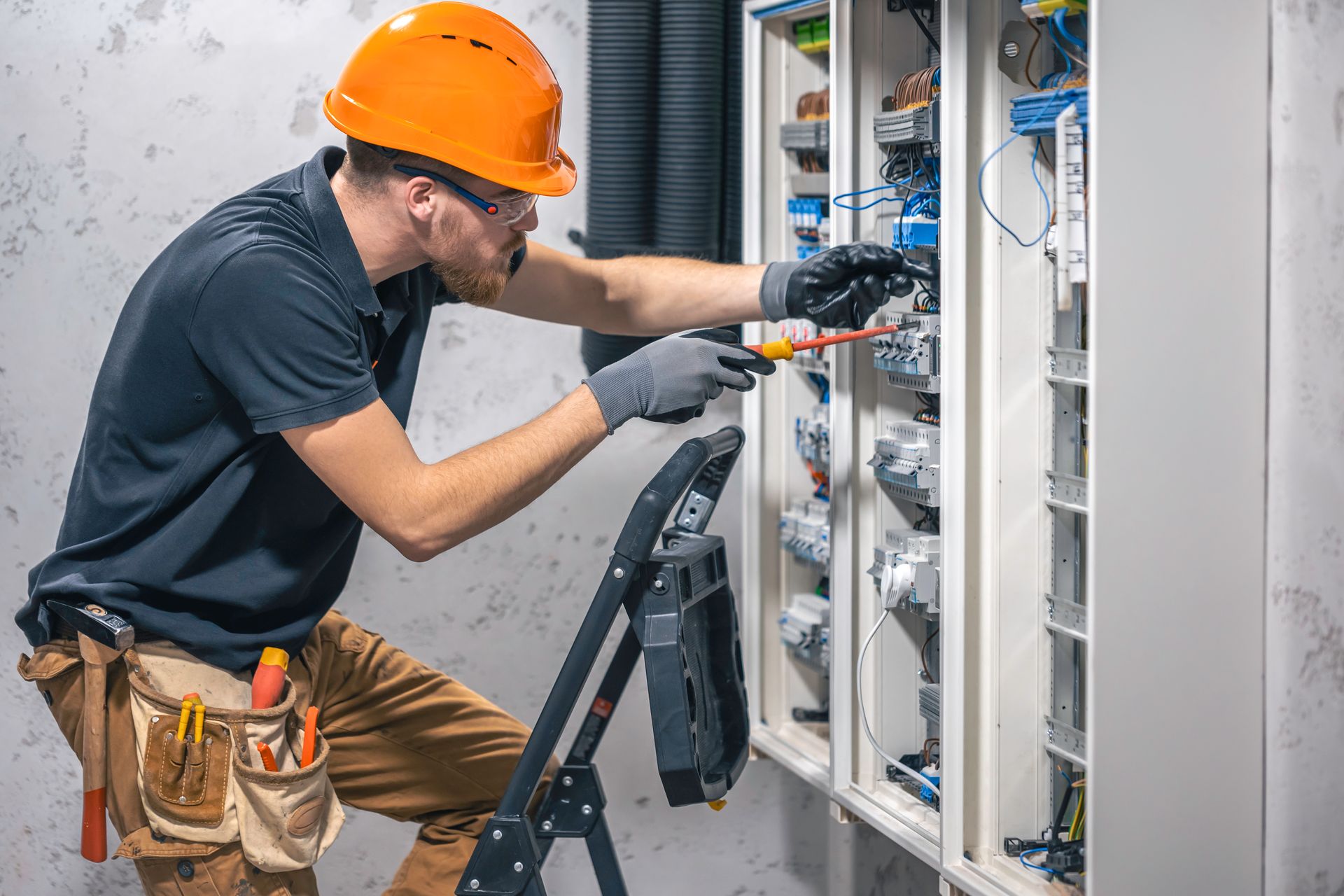 Electrician in orange hard hat working on an open electrical panel with tools on a ladder