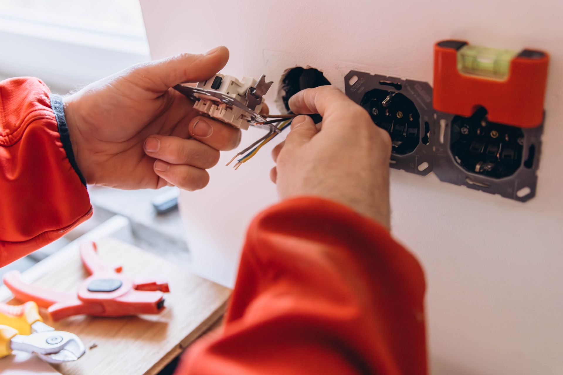 Hands wiring an electrical outlet in a wall with red sleeves visible